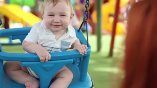 Egy kis vicces fiú műanyag hintákon egy külső játszótéren az anyjával. — Stock videók