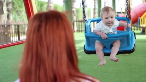 A vörös hajú nő megrázza a kisfiát műanyag hintákon. — Stock videók