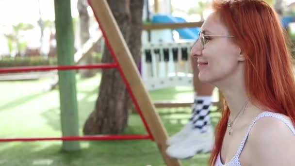 Roodharige vrouw swinging haar jonge zoon op plastic schommels — Stockvideo
