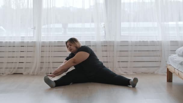 Şişman bir kadın yerde oturup esneme egzersizleri yapıyor. — Stok video