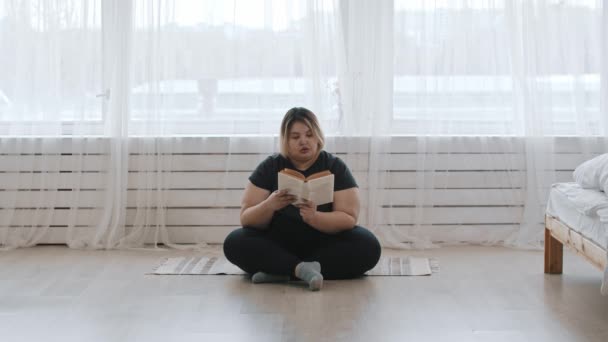 Una mujer gorda sentada en el suelo y leyendo un libro en casa — Vídeo de stock