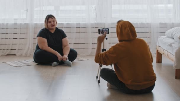 Selvsikker overvektig feminist og hennes støttende venn som sitter på gulvet og spiller inn en video for sine tilhengere – stockvideo