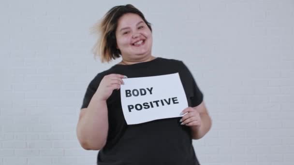 Concepto de positividad corporal - una mujer sonriente con sobrepeso bailando y sostiene un cartel con la inscripción CUERPO POSITIVO — Vídeo de stock
