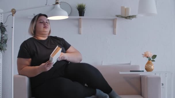 Una mujer gorda acostada en un sofá y leyendo un libro en casa — Vídeo de stock