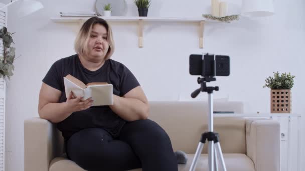Позитивність тіла - впевнена товста жінка-феміністка лежить на дивані і робить відео на своєму телефоні для її послідовників про позитивність тіла - читання цитат з книги — стокове відео