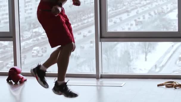 Muscular hombre sin camisa salta sobre la cuerda en el estudio con ventanas panorámicas guantes de boxeador en el suelo — Vídeos de Stock