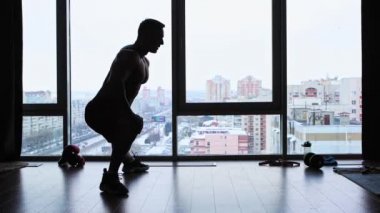 Stüdyoda panoramik pencereli spor eğitimi. Bacaklarını esneten üstsüz atletik bir adam silueti.