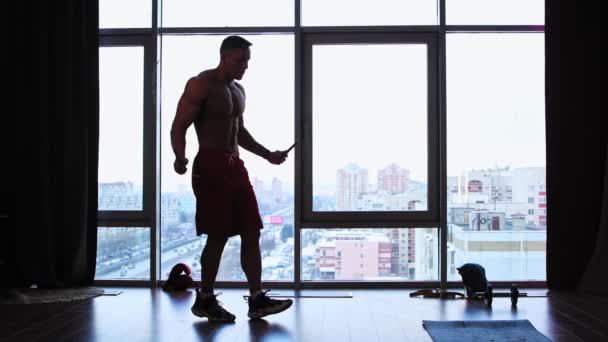 Entrenamiento deportivo en el estudio con ventanas panorámicas: una silueta de hombre atlético sin camisa saltando rápidamente sobre la cuerda — Vídeo de stock