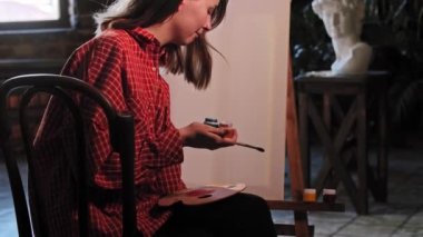 Sanat atölyesindeki genç bir kadın bir sehpanın önünde oturan renkli boyaları açıyor.