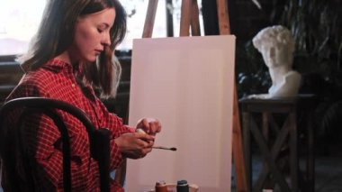 Sanat atölyesindeki genç bir kadın sehpanın önünde duran renkli boya kavanozlarını açıyor.