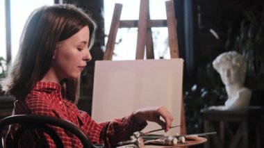 Sanat atölyesindeki genç bir kadın küçük bir fırça alıyor ve resim çizmek üzere.