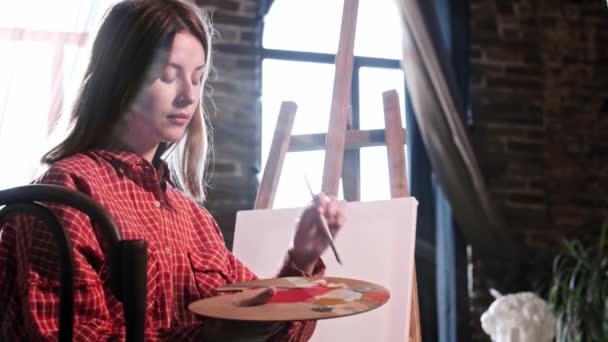 Μια νεαρή γυναίκα καλλιτέχνης ανάμειξη χρωμάτων στην παλέτα - κρατώντας ένα πινέλο στα δόντια της και βάζοντας τα μαλλιά της πίσω από τα αυτιά — Αρχείο Βίντεο
