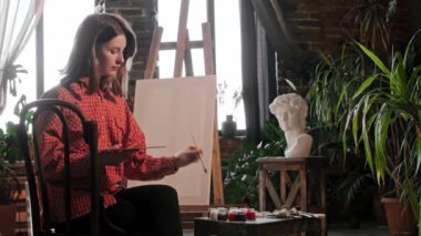 Genç ve güzel bir kadın sanatçı palette renkleri karıştırıyor bir sehpanın önünde oturuyor.