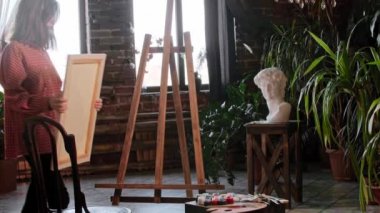 Genç ve güzel bir kadın ressam bir sehpaya tuval koyup önünde oturuyor.