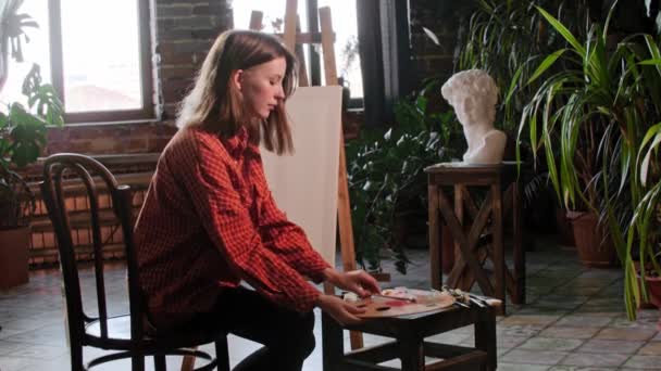 Μια νεαρή όμορφη γυναίκα καλλιτέχνης κάθεται μπροστά από ένα καβαλέτο και αρχίζει να αναμιγνύει χρώματα σε μια παλέτα — Αρχείο Βίντεο