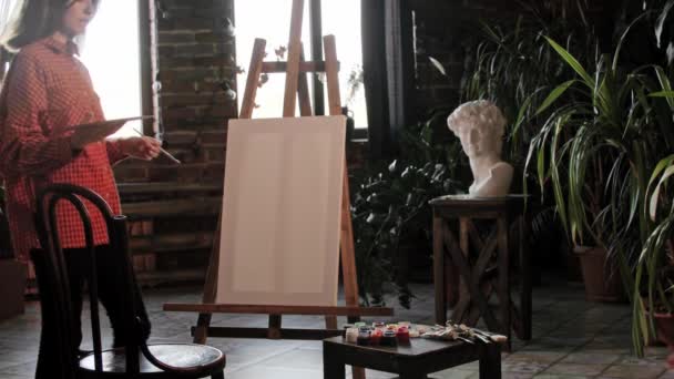 Μια νεαρή όμορφη γυναίκα καλλιτέχνης κάθεται μπροστά από ένα καβαλέτο και ανακατεύει χρώματα σε μια παλέτα — Αρχείο Βίντεο