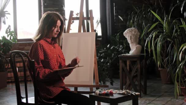 Μια νεαρή όμορφη γυναίκα καλλιτέχνης ανακατεύοντας χρώματα σε μια παλέτα και κοιτάζοντας τον άδειο καμβά — Αρχείο Βίντεο