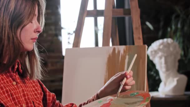 En ung söt kvinna artist tillämpa färger på en duk med en liten borste — Stockvideo