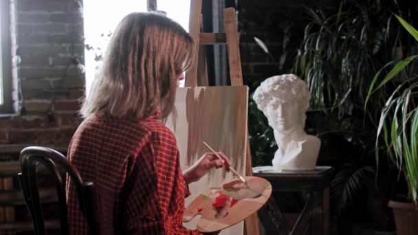 한 젊은 여성 미술가가 이젤 뒤를 보면서 붓으로 캔버스에 밑 색을칠 하고 있습니다 — 비디오