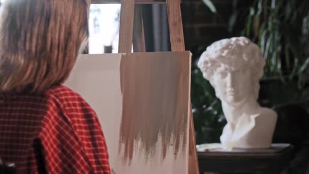 Une jeune femme artiste regardant derrière le chevalet et appliquant des couleurs de base sur une toile — Video
