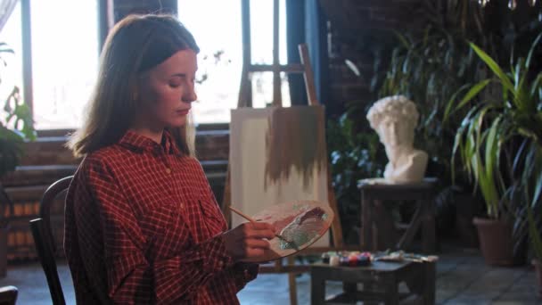En ung kvinnlig konstnär blandar färger på en träpalett och tittar in i kameran — Stockvideo