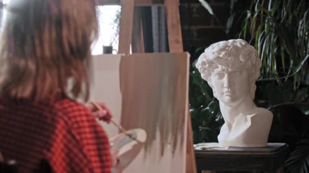 Μια νεαρή γυναίκα καλλιτέχνης που κοιτάζει ένα ελληνικό κεφάλι γλυπτό και ανάμειξη χρωμάτων σε μια παλέτα — Αρχείο Βίντεο