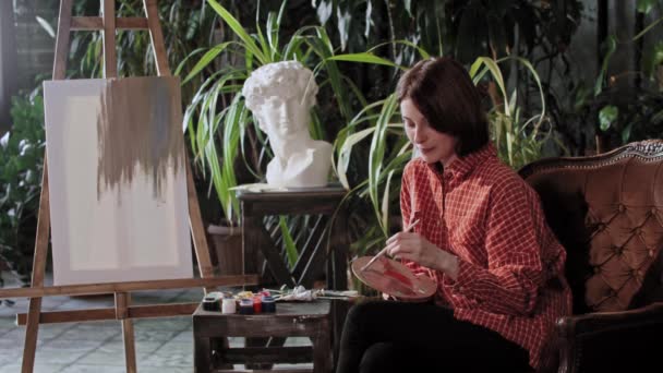 Mladá krásná žena umělec míchání barev na dřevěné paletě sedí na židli - dívá se do kamery — Stock video
