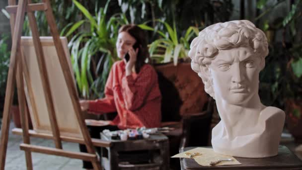 Artista em um estúdio de arte com plantas - jovem mulher bonita fala em seu telefone sentado na frente de um cavalete - escultura cabeça grega em primeiro plano — Vídeo de Stock