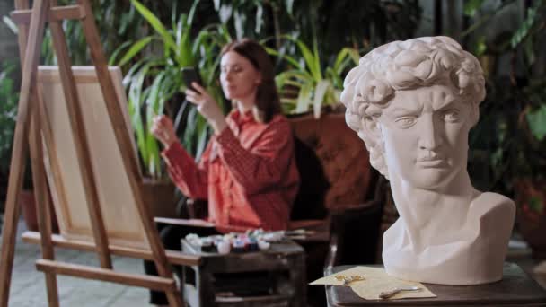 Artista em um estúdio de arte com plantas - jovem mulher bonita tira uma foto de sua pintura - escultura cabeça grega em primeiro plano — Vídeo de Stock
