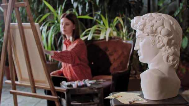 Kunstenaar in een atelier met planten - jonge mooie vrouw tekent een grieks hoofd sculptuur op de voorgrond — Stockvideo