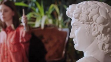 Bitkilerle dolu bir sanat stüdyosunda sanatçı. Genç, güzel bir kadın tuvale Yunan heykeli çiziyor.