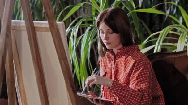 Kunstenaar in een atelier - jonge mooie vrouw tekening van een schilderij met kleuren en op zoek naar de verwijzing naar de zijkant — Stockvideo