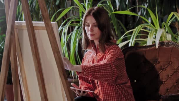 Artista en un estudio de arte con plantas - joven mujer bonita dibujando una pintura con colores — Vídeos de Stock