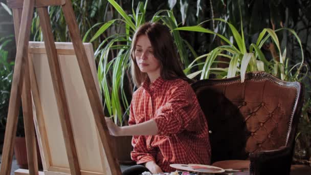 Konstnär i en konststudio med växter - ung vacker kvinna ritar en målning på ett staffli och sträcker sig sedan upp med ett leende på läpparna — Stockvideo
