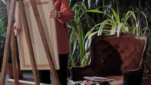 Kunstenaar in een atelier met planten - jonge mooie vrouw brengt een doek in de studio en zet het op een ezel — Stockvideo