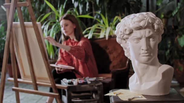 El artista en el estudio de arte con las plantas - la mujer joven bonita que dibuja la pintura - la escultura griega de cabeza en el primer plano — Vídeos de Stock
