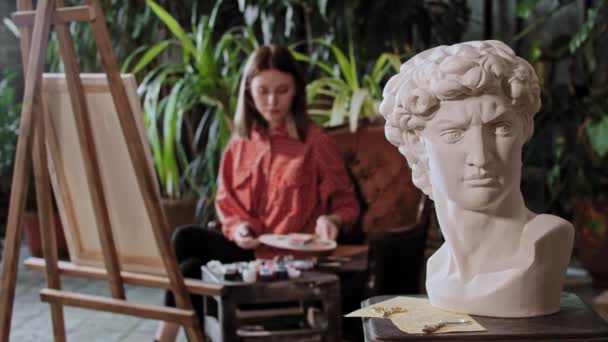 Artiste dans un atelier d'art avec des plantes - jeune femme artiste mettre la toile loin d'un chevalet - sculpture de la tête grecque sur un premier plan — Video