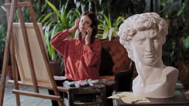 Artista em um estúdio de arte com plantas - jovem mulher bonita fala em seu telefone - escultura cabeça grega em primeiro plano — Vídeo de Stock