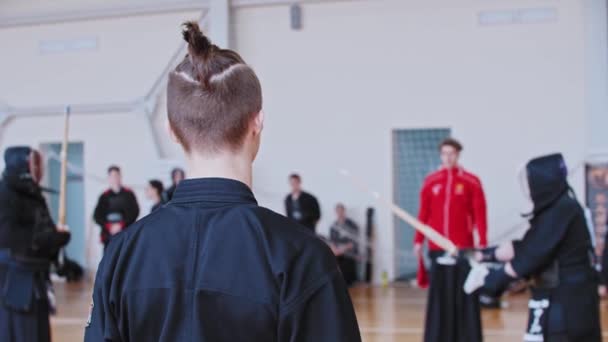 Turniej Kendo - ludzie walczą na siłowni - człowiek z ogolonymi świątyniami i wysokim kucykiem oglądającym walkę — Wideo stockowe