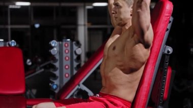 Formda bir adam egzersiz aletinin üzerinde oturuyor ve halterleri kaldırmaya başlıyor.