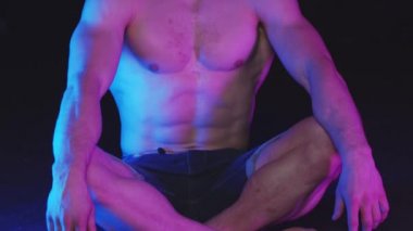 Kot şortlu genç adam neon ışıklı siyah stüdyoda oturuyor.