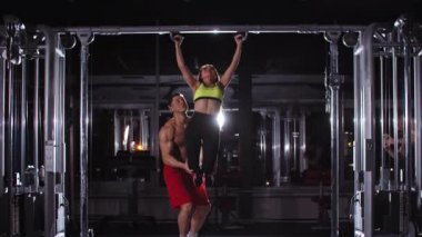 Spor salonunda kadın ve erkek antrenman yapıyor. Bir adam, kadının dizlerinden tutup barfiks çekmesine yardım ediyor.