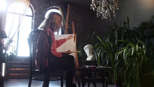 Een jonge mooie vrouw zit in de kunstwerkplaats en brengt bruine tinten aan op een doek — Stockvideo