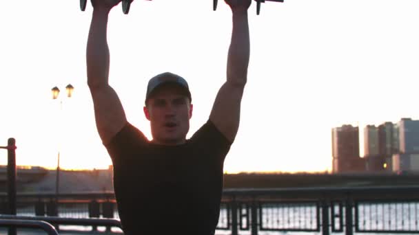 Молодий спортсмен тренує руки гантелями на свіжому повітрі на заході сонця — стокове відео