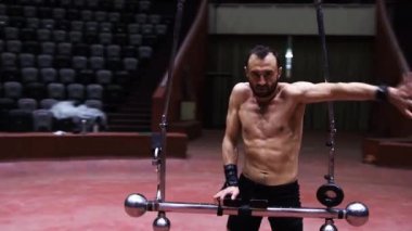 Sirkin uçuş çubuğunda performans gösteren akrobatik bir adam bir eliyle bara yaslanarak ayağa kalkar.
