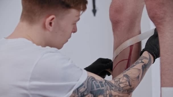 Jongeman tatoeage kunstenaar plakken een duct tape op het been op zijn cliënt — Stockvideo