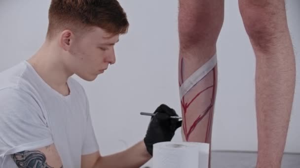 Jongeman tatoeage kunstenaar tekening een schets op het been op zijn cliënt met een marker — Stockvideo