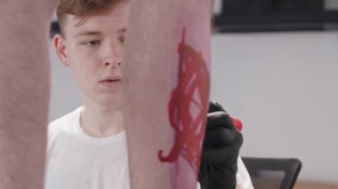 Genç bir dövme sanatçısı, elinde kırmızı bir kalemle müvekkilinin bacağına soyut bir çizim yapıyor.