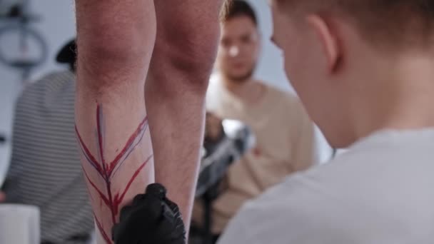 若い男の入れ墨のアーティストは、マーカーの上にペンで彼のクライアント上の足の上にスケッチを描く — ストック動画