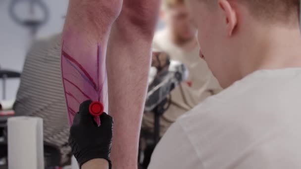 Joven tatuador dibujando un boceto en la pierna de su cliente con un marcador rojo — Vídeos de Stock
